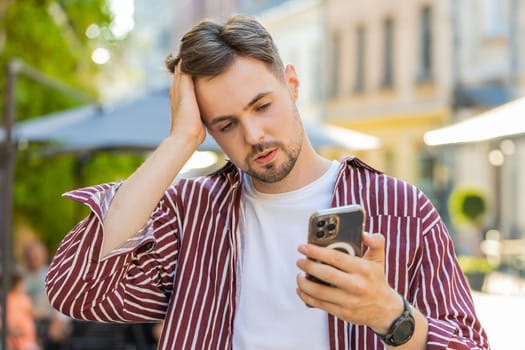 Bearded Caucasian man use smartphone typing browsing, loses becoming surprised sudden lottery results, bad news, fortune loss, fail. Young guy walking in urban city street outdoors. Town lifestyles