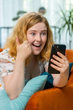 Happy woman use mobile smartphone typing browsing say Wow yes found out great big win good news celebrate lottery jackpot, play victory game. Redhead girl at home in room lying on sofa. Vertical shot