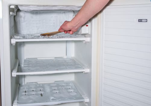 ice has formed on the walls of the freezer and it is creeping out, a woman's hand is knocking the ice off with a wooden spatula,