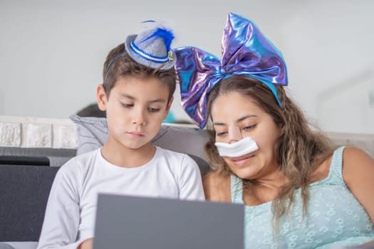 Smiling mother with bondage on her face after rhinoplasty lies in bed with her little son and watches movie on digital tablet,concept of joint leisure