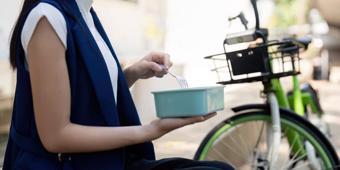 A friendly businesswoman holds a bowl with a fork, smiling while seated next to her sustainable bicycle in a bright outdoor setting, promoting a healthy and eco conscious lifestyle.