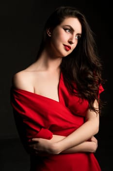 Amazing young bright brunette woman in red dress with bare shoulder standing with crossed arms and looking along