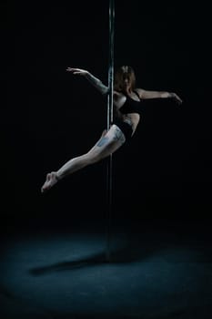Young flexible woman with tattoos posing on pylon with outstretched arms on black background