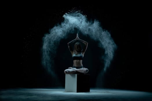 Young flexible woman in lingerie posing on cube while performing in cloud of white dust