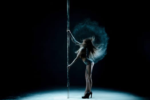 Graceful girl wearing lingerie and high heels dancing on pylon in dust cloud view in the dark room