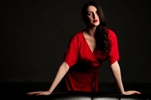 Sexy brunette in red dress leaning against black leather sofa shot in the dark room