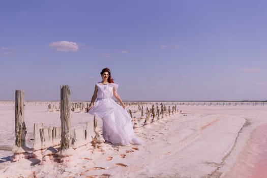 A woman in a white dress walks on a pink salt flat, passing wooden posts, against a bright blue sky.