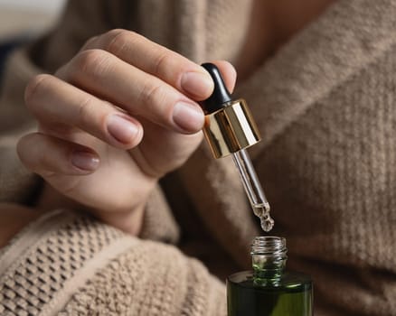 Female hands with natural manicure opening white green cosmetic dropper bottle. Close up of woman in bathroom wearing light brown bathrobe with drop of serum or essence falling from pipette