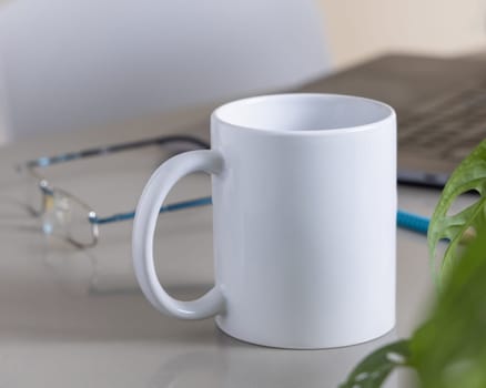 Closeup of 11 oz white blank coffee mug on grey office table against lopened laptop, glasses and green plant, mockup. Template for design with copy space