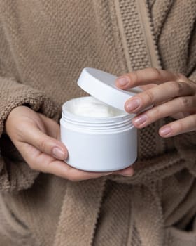 Female hands with natural manicure opening white blank cream jar, cosmetic mockup. Close up of woman in bathroom wearing light brown bathrobe. Bodycare concept