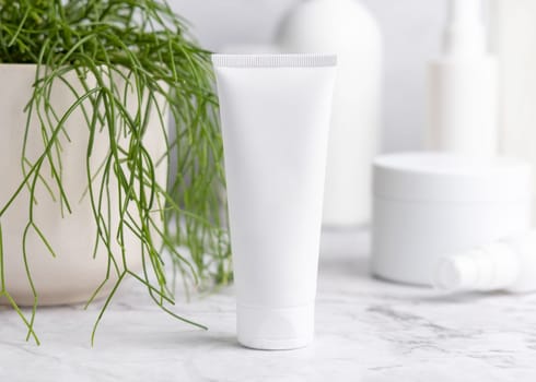 White cream squeeze tube on white marble table against green plant and cosmetic bottles in bathroom, close up, packaging mockup. Natural product for face care