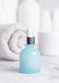Light blue glass dropper bottle on white marble against towel and cosmetics in bathroom, close up, packaging mockup. Natural product for facecare - serum or essence