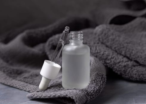 Pipette set on opened clear glass dropper bottle near dark grey bath towel closeup, brand packaging mockup. Natural handmade cosmetic product for facecare and spa procedures - serum or essence