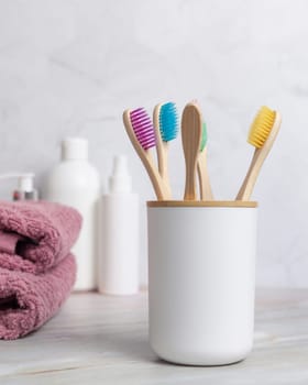Eco-friendly colorful bamboo toothbrushes in holder cup in a white in bathroom against natural cosmetics and towel, mockup. Personal care and hygiene products