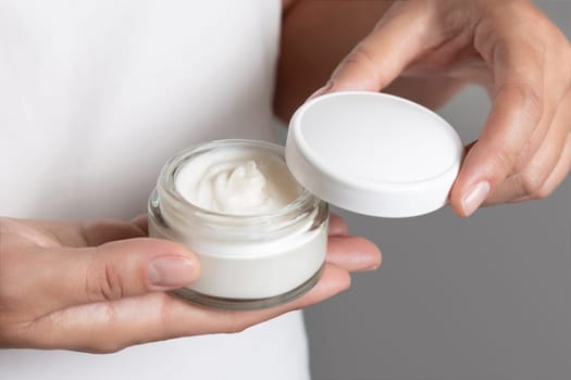 Woman opening cream jar against white shirt and grey background, fingers with natural manicure, cosmetic mockup. Concept of organic cosmetics for body or face care