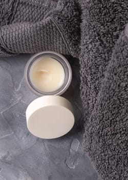Opened lip balm jar with blank lid near dark grey folded towel top view, cosmetic packaging mockup, copy space. Lipcare beauty product for mens everyday personal care