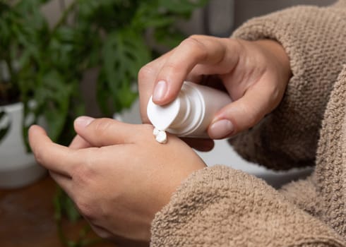 Close up of woman in brown bathrobe applying cream on hand from a white cosmetic bottle in bathroom against green plant and vessel basin. Organic cosmetics for daily handcare