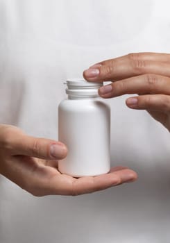 Closeup of woman in white t-shirt holding a white plastic medical pill bottle. Packaging mockup for herbal supplement. Female hands with natural manicure