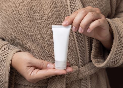 Close up of woman in light brown bathrobe with white blank cream tube in hands, mockup. Hands with natural manicure holding squeeze cosmetic tube - lotion or shampoo