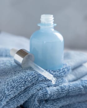 Drop falling from pipette put near opened blue glass dropper bottle on blue towel against grey wall close up, mockup. Natural serum or essence