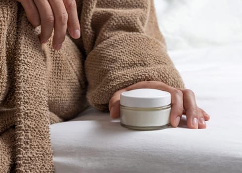 Woman in brown bathrobe puts white opened cream jar on white bedsheet closeup. Bodycare concept. Organic cosmetics for body and face care, beauty and spa procedures