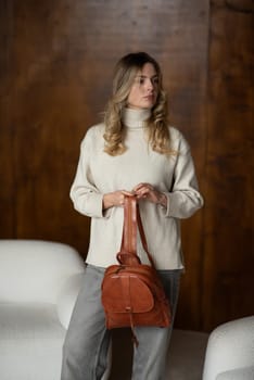 beautiful girl in gray pants and beige sweater posing with a small brown leather backpack.