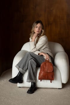 beautiful girl in gray pants and beige sweater posing with a small brown leather backpack.
