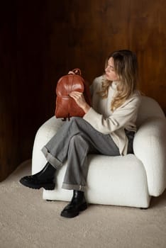 beautiful girl in gray pants and beige sweater posing with a small brown leather backpack.