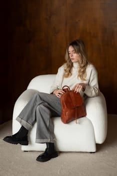 beautiful girl in gray pants and beige sweater posing with a small brown leather backpack.
