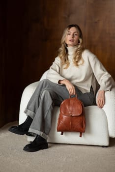 beautiful girl in gray pants and beige sweater posing with a small brown leather backpack.