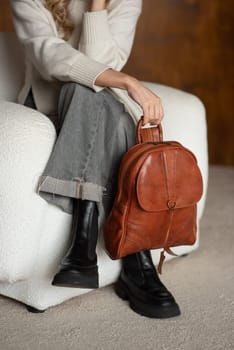 beautiful girl in gray pants and beige sweater posing with a small brown leather backpack.