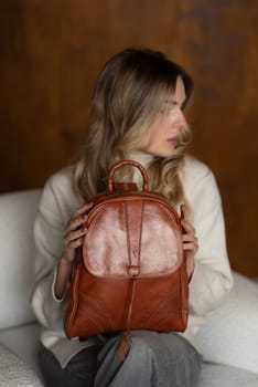 beautiful girl in gray pants and beige sweater posing with a small brown leather backpack.