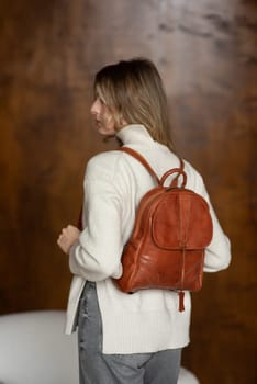 beautiful girl in gray pants and beige sweater posing with a small brown leather backpack.