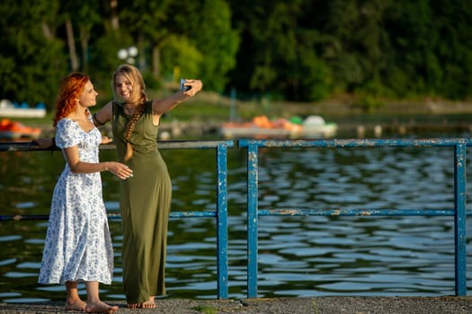 Two close friends happily take a selfie by the shimmering water, capturing a truly joyful summer moment together