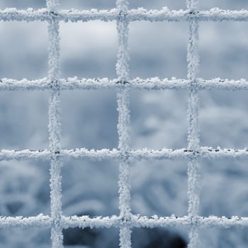 A minimalist close-up of a frosted metal grid, delicately coated in ice crystals, set against a soft, blurred winter background. The intricate patterns of the frost highlight the geometric symmetry of the grid, creating a serene and abstract winter composition.