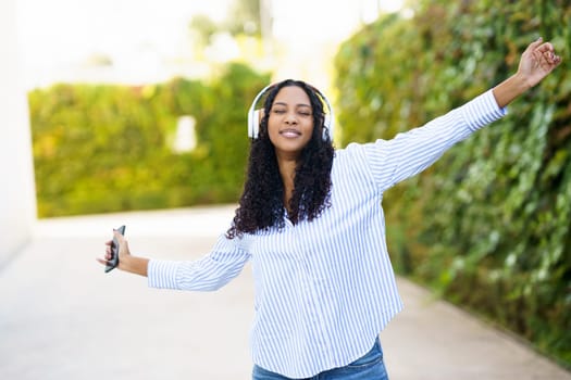A cheerful young woman dances joyfully outdoors in a stunning vibrant green setting, donning stylish headphones while immersing herself in the rhythm of her favorite music and feeling free