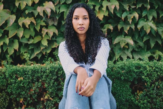 A young woman with beautifully curly hair sits thoughtfully in front of a lush, vibrant green foliage, showcasing a serene and peaceful expression that reflects her calm state of mind