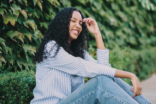 A cheerful and vibrant young woman is seated comfortably against a lush green backdrop, wearing a stylish striped shirt and smiling joyfully, truly embodying happiness and positivity in her presence