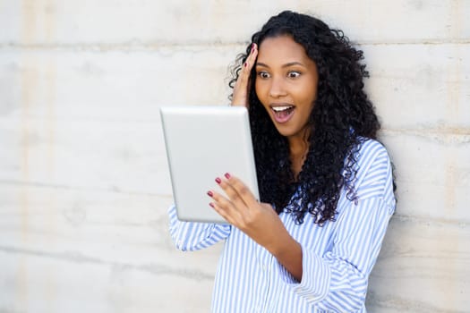 A young woman with beautifully curly hair radiates surprise and beams with joy while enthusiastically using a tablet, showcasing a lively moment filled with genuine engagement and deep emotion