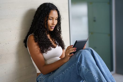 A young woman with beautifully styled curly hair sits comfortably against a chic wall, fully engrossed in her tablet, enjoying a joyful and peaceful moment of leisure and modern technology