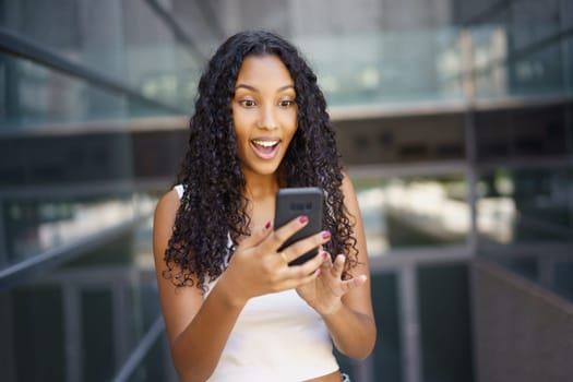 A young woman with beautiful curly hair is expressing her excitement and joy, while happily using her smartphone in a vibrant and contemporary environment filled with modern dynamics and energy