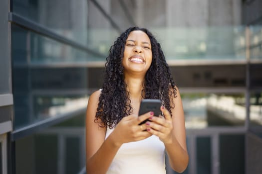 A cheerful and vibrant young woman holds her smartphone up while smiling widely, fully enjoying a lovely sunny day in a lively urban setting filled with energy and excitement all around her