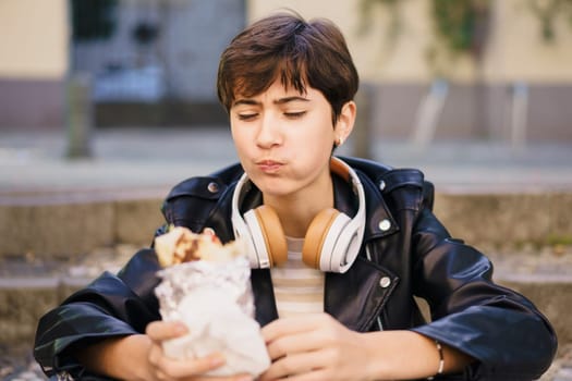 A young individual is sitting comfortably outdoors, fully enjoying a delicious and flavorful wrap while wearing headphones, surrounded by the lively atmosphere of a vibrant urban city