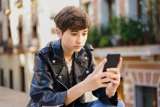 In a vibrant and lively urban area, a young girl dressed in a stylish leather jacket is sitting outdoors, fully absorbed in her smartphone, surrounded by the bustling energy of the city itself