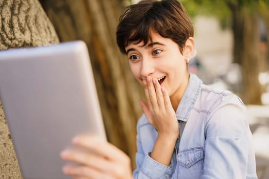 A young woman with a stylish short haircut displays a mixture of surprise and genuine joy as she gazes at her tablet, fully capturing authentic emotions while enjoying a relaxed outdoor setting