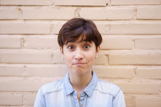 A vibrant young woman with a stylish short haircut shows surprise as she confidently poses against a lightbrick wall, embodying the emotion and diversity of contemporary urban life