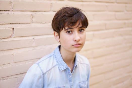 A confident and creative young individual, sporting short hair, stands proudly in front of a beige wall wearing a stylish denim shirt, which highlights their relaxed yet trendy and fashionable style