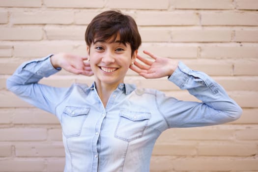 A joyful, vibrant young woman with short hair smiles and poses playfully against a light brick wall, radiating warmth, positivity, and confidence with her cheerful demeanor and carefree attitude