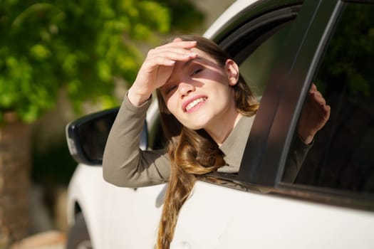 A cheerful young woman, happily leans out of a car window while playfully shielding her eyes from the bright sun on a beautiful and sunny day filled with adventure and joy