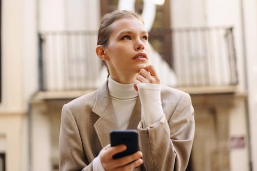 In a charming urban setting, a young woman in an elegant coat and cozy turtleneck focuses on her smartphone, lost in thoughts while enjoying her beautiful surroundings with a sense of peace